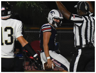 A person in a football uniform holds a football while a referee signals with raised arms and another player stands nearby on a nighttime field. Description generated by AI