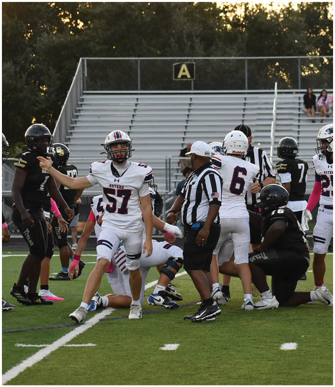People in football uniforms and referees are gathered on a field during a game with empty bleachers and a few spectators in the background. Description generated by AI