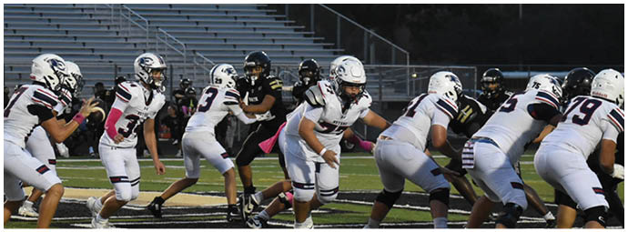 A group of people wearing football uniforms are lined up on a field preparing for a play during a game at dusk. Description generated by AI