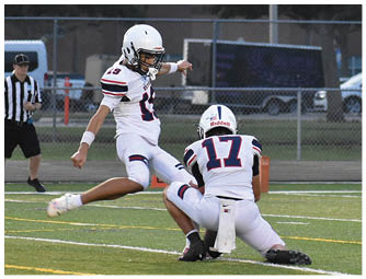 Two people in football uniforms are on a field with one holding the ball on the ground while the other is kicking it. Description generated by AI