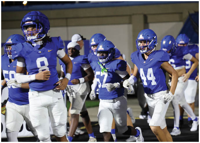 Three football players in blue and white uniforms running on a field. Description generated by AI