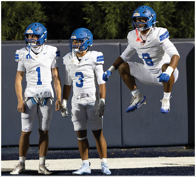 Three football players in blue and white uniforms running on a field. Description generated by AI