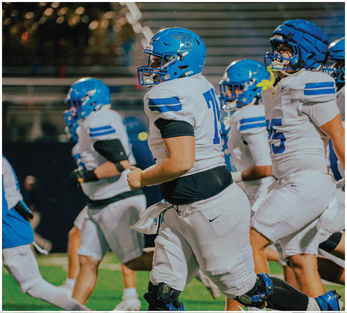 Three football players in blue and white uniforms running on a field. Description generated by AI