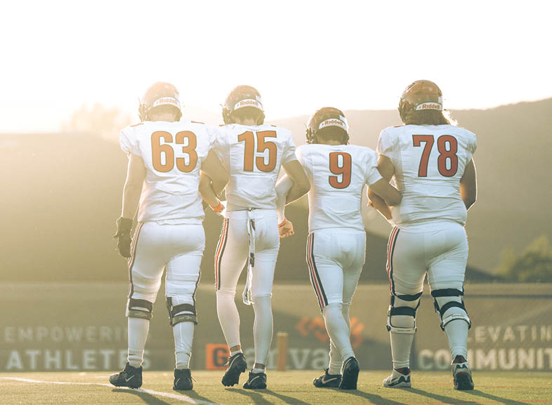 Four people wearing black football uniforms with “VISTA" on the front are standing together in a stadium with a crowd in the background, two of them holding helmets raised above their heads. Description generated by AI