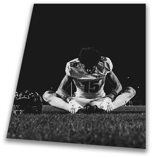 A person in a black football uniform and helmet is crouched in a ready position on a green field with another player in white uniform visible in the background. Description generated by AI