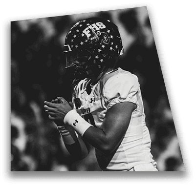 A person in football gear and helmet is crouched on a field with other players lined up in the background during a game. Description generated by AI