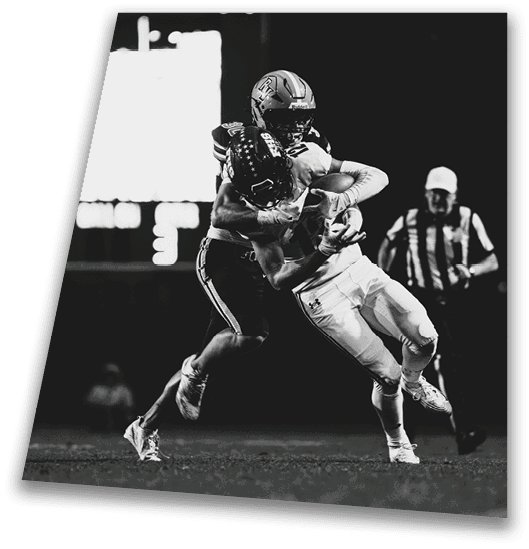 A person in a black football uniform and helmet is crouched in a ready position on a green field with another player in white uniform visible in the background. Description generated by AI