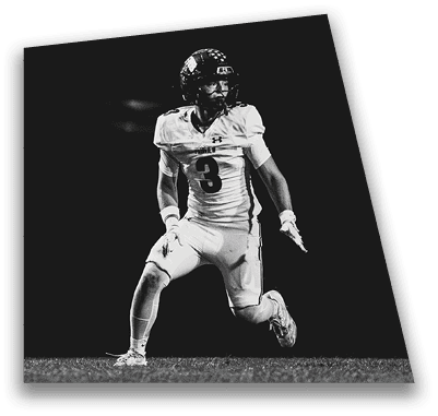A person in football gear and helmet is crouched on a field with other players lined up in the background during a game. Description generated by AI