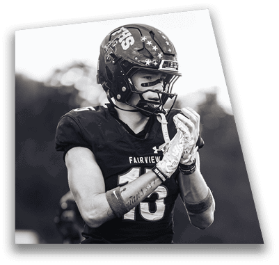 A person in football gear and helmet is crouched on a field with other players lined up in the background during a game. Description generated by AI