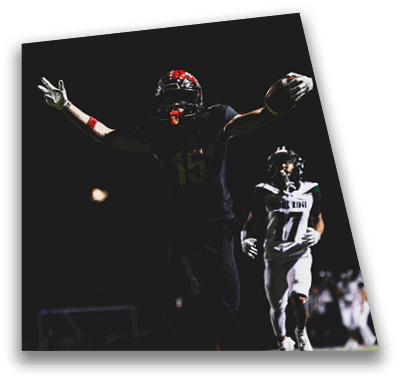 A person in football gear and helmet is crouched on a field with other players lined up in the background during a game. Description generated by AI