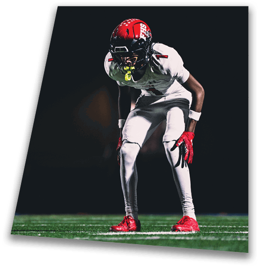 A person in a black football uniform and helmet is crouched in a ready position on a green field with another player in white uniform visible in the background. Description generated by AI