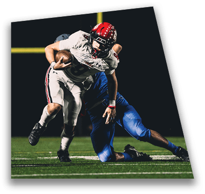 A person in football gear and helmet is crouched on a field with other players lined up in the background during a game. Description generated by AI
