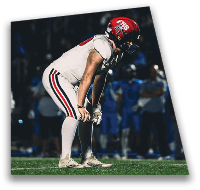 A person in football gear and helmet is crouched on a field with other players lined up in the background during a game. Description generated by AI
