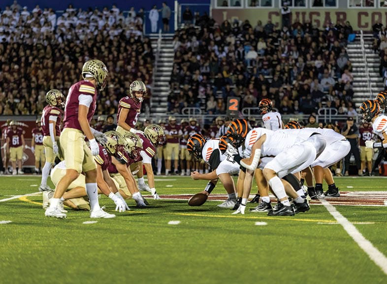 Four people wearing black football uniforms with “VISTA" on the front are standing together in a stadium with a crowd in the background, two of them holding helmets raised above their heads. Description generated by AI