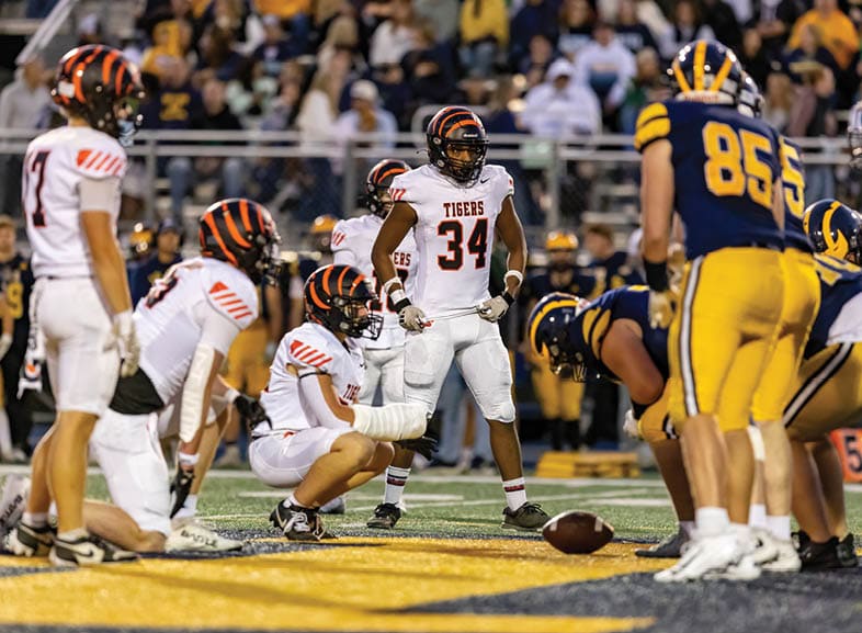 Four people wearing black football uniforms with “VISTA" on the front are standing together in a stadium with a crowd in the background, two of them holding helmets raised above their heads. Description generated by AI