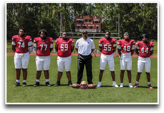 Seven people wearing red football jerseys and white pants stand side by side on a football field with trees and a scoreboard in the background. Description generated by AI