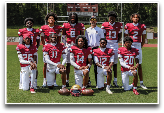 A group of people wearing red and white football uniforms pose on a grassy field with helmets and footballs placed in front of them. Description generated by AI