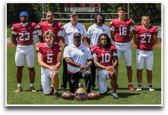 Eight people wearing football uniforms and two people in white polo shirts pose together on a football field with a scoreboard and trees in the background. Description generated by AI