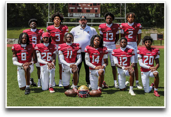 A group of football players in red and white uniforms and a coach in a white shirt pose on a grassy field with footballs and helmets in front of them. Description generated by AI