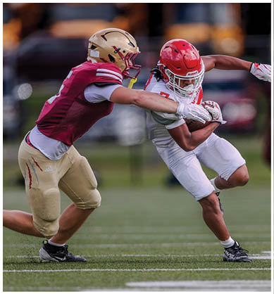 Two people in football uniforms are actively engaged in a play on a grassy field with one person attempting to tackle the other who is holding the ball. Description generated by AI
