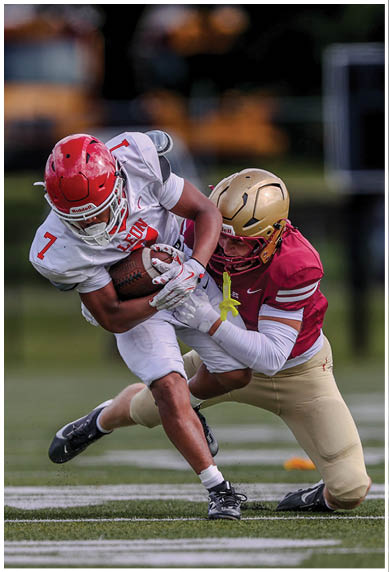 A person in a white football uniform with a red helmet is running with the ball while another person in a red and gold uniform is tackling them on a football field. Description generated by AI