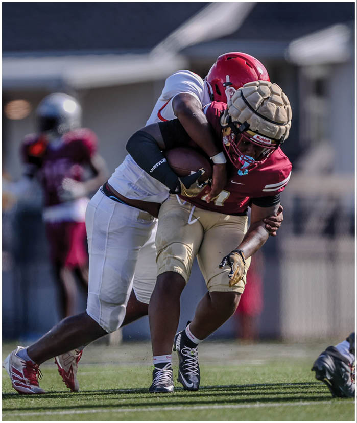 Players in red and white football uniforms are engaged in a tackle on a grassy field during a game. Description generated by AI