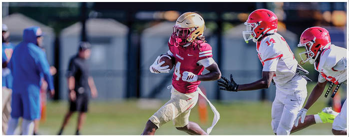 An adult in a red sports uniform is running with a football while two adults in white uniforms chase behind on a grassy field. Description generated by AI