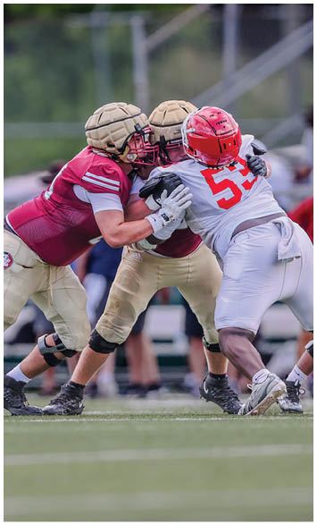 Two people in maroon and gold football uniforms are blocking a person in a white and red football uniform during a game on a grassy field. Description generated by AI