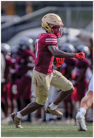 A person in a maroon and gold football uniform is running on a field toward another person in a white and red football uniform with a blurred crowd and school bus in the background. Description generated by AI