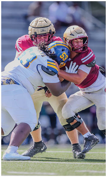 Three people wearing football helmets and uniforms are engaged in a close physical play on a grassy field during a game. Description generated by AI