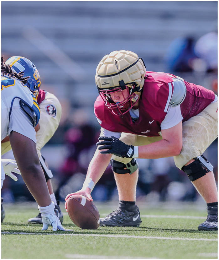 A person in football gear is crouched on a field holding a football, preparing for a play against an opposing player. Description generated by AI