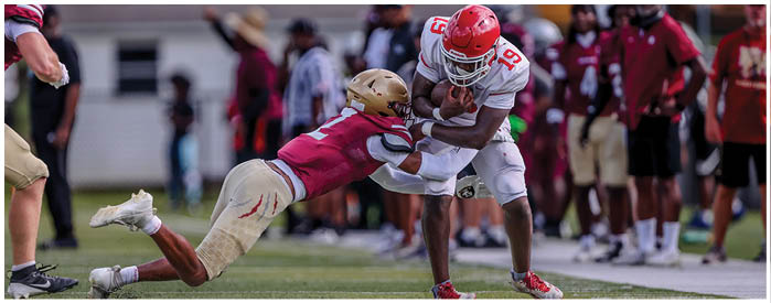 Two people in football uniforms are engaged in a tackle on a grassy field with others and a fence in the background. Description generated by AI