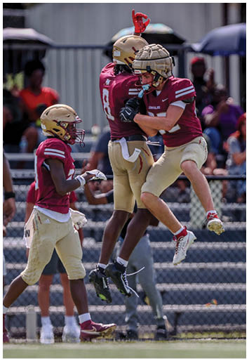 Three football players in maroon and gold uniforms celebrate on the field with one player jumping and another raising a hand while spectators watch from the stands. Description generated by AI