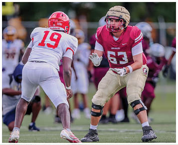 Two people in football uniforms are facing each other on a field with one in a red jersey and the other in a maroon jersey preparing for a play. Description generated by AI