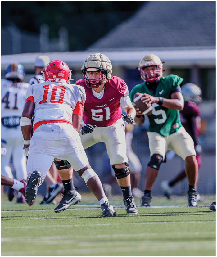Several people wearing football uniforms and helmets are actively engaged in a play on a grassy field with a blurred background of a building and fencing. Description generated by AI
