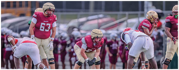 Several people wearing football helmets and uniforms are positioned on a field, preparing for a play during a game. Description generated by AI