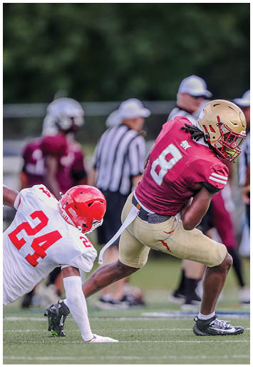A person wearing a maroon football jersey with the number 8 and a gold helmet is running with the ball while another person in a white jersey and red helmet attempts to tackle them on a football field. Description generated by AI