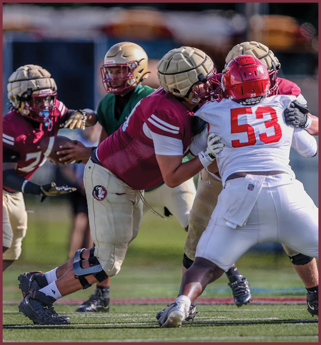 Several football players in helmets and uniforms are engaged in a play on a grassy field with school buses and a building in the background. Description generated by AI