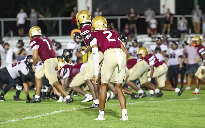 Several people wearing maroon and gold football uniforms are positioned on a grassy field preparing for a play against opponents in white and black uniforms during a nighttime game. Description generated by AI