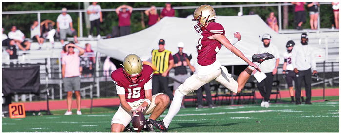 Two people in football uniforms are on a field with one kicking a ball while the other bends forward, with spectators and officials in the background. Description generated by AI