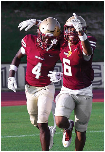 Two people wearing maroon football uniforms and helmets celebrate on a grassy field with one person pointing forward and the other with an arm around their shoulder. Description generated by AI