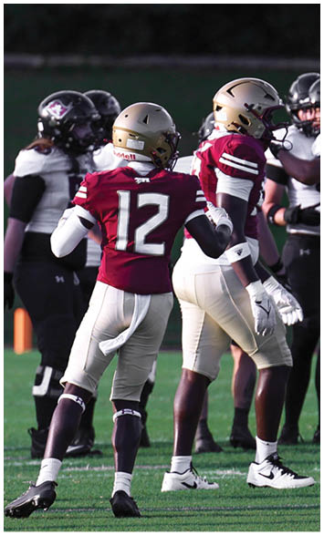 A person wearing a maroon football jersey with the number 12 and a gold helmet is on a football field with other players in the background. Description generated by AI