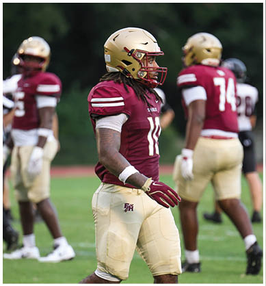 A person wearing a maroon and gold football uniform and helmet is walking on a grassy field with other players and a referee in the background. Description generated by AI