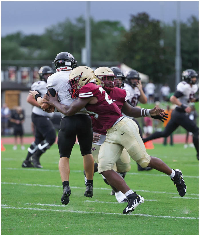 Two people in football uniforms are engaged in a tackle on a grassy field during a game with other players and spectators in the background. Description generated by AI