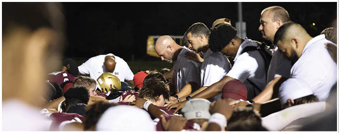 A group of people wearing sports attire are gathered closely with heads bowed and hands placed on each other in a moment of prayer or reflection on a field at night. Description generated by AI
