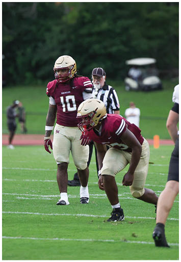Two people in maroon and beige football uniforms are on a grassy field with one person standing and the other in a crouched position while a referee and other individuals are visible in the background. Description generated by AI