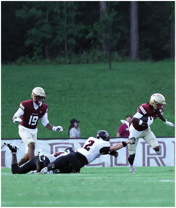 A person in a maroon and white football uniform is running while another person in a white and black uniform is diving and reaching out to try to tackle them on a grassy field. Description generated by AI