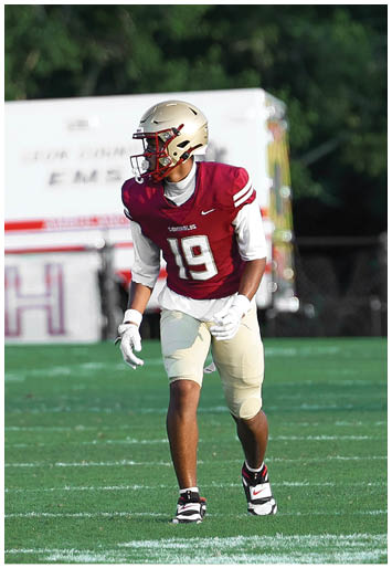 A person wearing a gold football helmet and a red jersey with white stripes is looking to the side outdoors with trees in the background. Description generated by AI