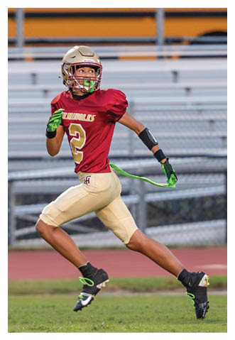 A person wearing a maroon and gold football uniform with the number 2 is running on a grassy field with a green flag football belt while looking to the side. Description generated by AI