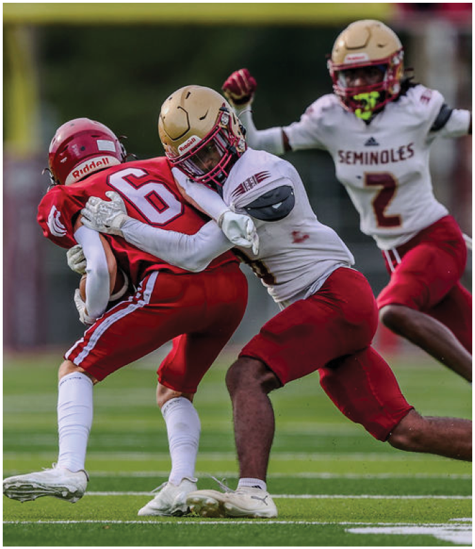 Four people in football uniforms are on a field with two players in white and red tackling a player in red while another player in white and red runs behind them. Description generated by AI